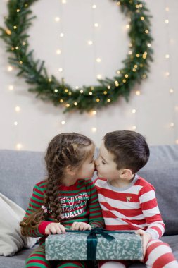 Anaokulu çocuğu ve kız Noel pijamalarıyla ellerinde kurdeleyle Noel çelengi altında ışıklarla öpüşüyorlar.