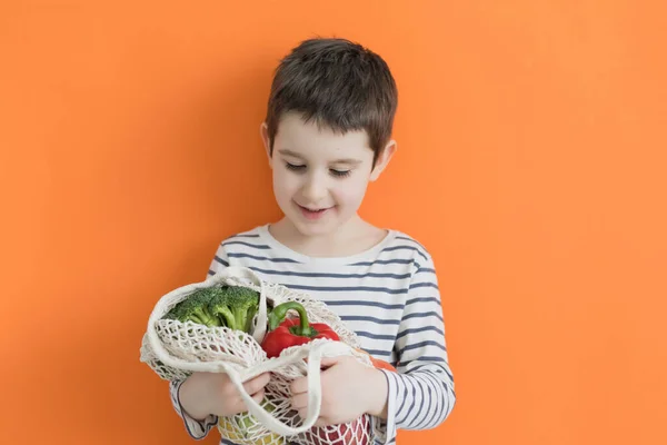 Child holds eco bag with fresh vegetables from farm market on an orange background with copy space. Healthy food concept, fresh vegetables, farm market