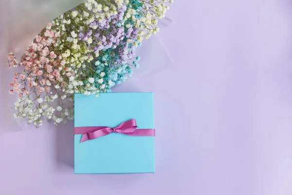 Rainbow gypsophila bouquet in package with ribbons and blue gift box on a violet background with copy space. Holiday gift concept