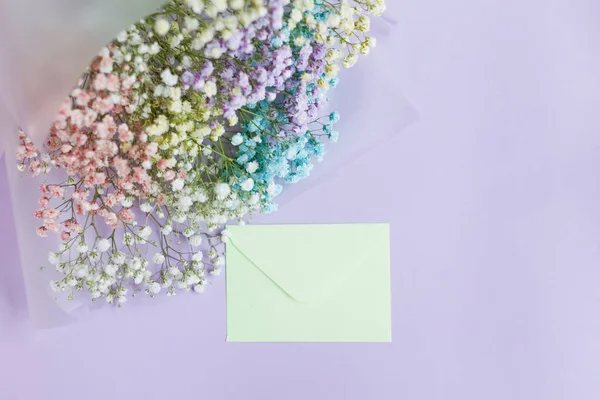 Rainbow gypsophila bouquet in package with ribbons and green envelope on a violet background with copy space. Holiday gift concept