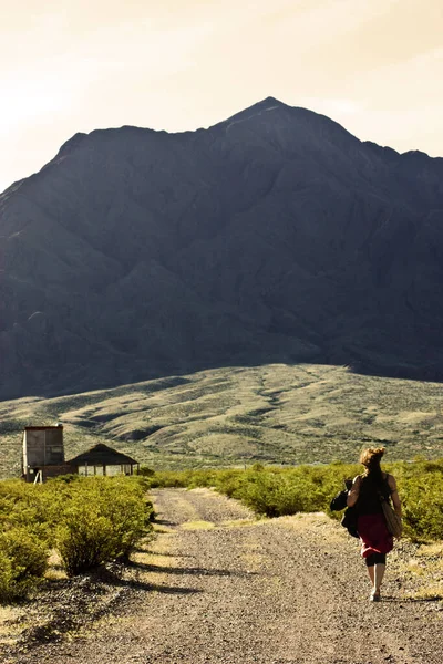 Sırtı kırmızı etekli bir kadın dağın yanındaki eve doğru toprak yolda yürüyor..