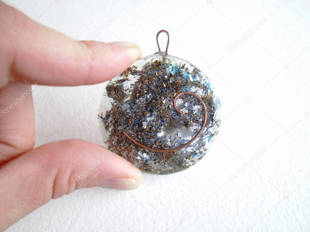 Orgone pendant made of transparent resin with auric copper spiral and the Hindu symbol of the 5th chakra Vishuddha held by a female caucasian hand. Harmonizer of energy fields. Ion and proton stabilizer.