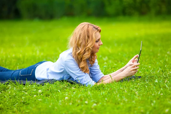 Mujer rubia acostada en hierba verde con una tableta en las manos —  Fotos de Stock