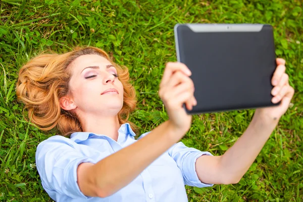 Mujer rubia bastante joven acostada en la hierba usando una tableta —  Fotos de Stock