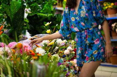 Çiçekçi dükkanının çiçeklerine ve bitkilerine, yaz elbisesi giyen bir kadınla seçici bir şekilde odaklan.