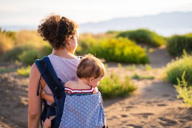 Bir annenin sırtında kumlu bir yolda bir bebeği sırtında taşırken yürümesi.