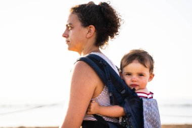 Bir bebek taşıyıcısının sırtında bebek taşırken ayakta duran bir annenin profili.