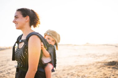 Anne, gün batımında bir kumsalda, sırtında bir keple bir bebek taşıyacak.