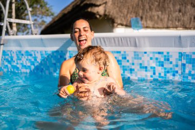 Beyaz bir kadın yazın havuzda bebeğiyle oynarken gülüyor.