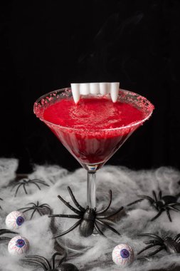 Cadılar Bayramı için vişne kokteylinin üst görüntüsü vampir dişleriyle, masada örümcekler ve gözlerle, seçici odak, siyah arka plan, dikey