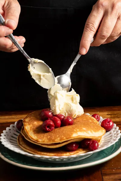 Top view of woman\'s hands with spoons and mascarpone cheese on pancakes with cherries, portrait