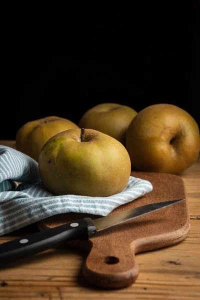 Top view of four pippin apples on wooden board, knife and rag on rustic wooden table, selective focus, black background, vertical