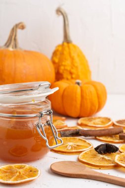 Kurumuş portakal dilimleri ve turuncu balkabaklarıyla beyaz masada balkabağı reçeli olan cam kavanozun yakın plan çekimi, beyaz arka plan, dikey