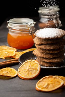 Tereyağlı kurabiyelerin üst görüntüsü, koyu renkli masaya düşen şeker, portakal dilimleri ve arka planda bir kavanoz reçel, seçici odak, dikey