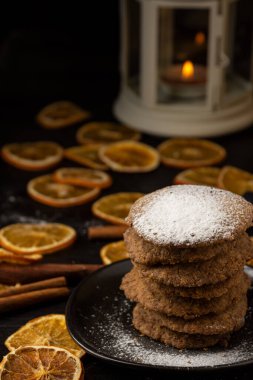 Koyu renkli masada şekerli kurabiye, turuncu dilimler, tarçın çubukları ve yanan mumlu beyaz fener, seçici odak, dikey