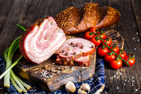 smoked pork meat, spiced bacon, slices, fresh vegetables, herbs and spices, on a black wooden, cutting kitchen board, rustic style dark and moody, black background