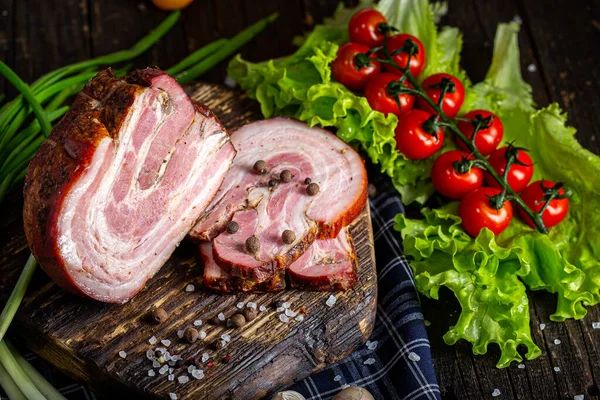 smoked pork meat, spiced bacon, slices, fresh vegetables, herbs and spices, on a black wooden, cutting kitchen board, rustic style dark and moody, black background