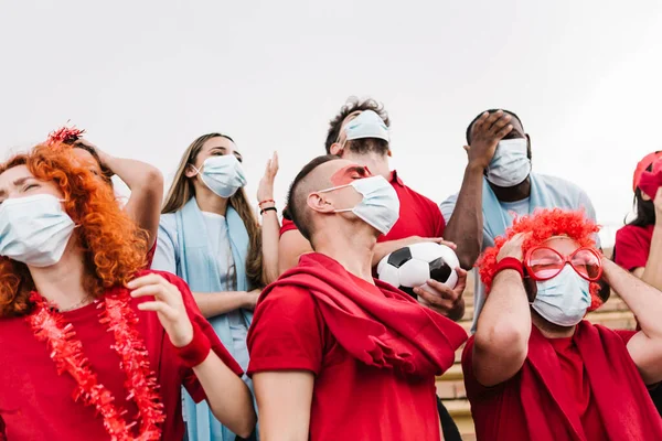 Gefrustreerde groep multiraciale voetbalfans in beschermende gezichtsmasker kijken naar hun voetbalteam verliezen - Focus op de mens in het centrum — Stockfoto
