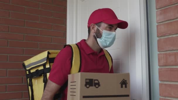 Messager hispanique en masque protecteur, capuchon rouge et t-shirt avec sac à dos thermique sonnant la sonnette de la porte pour livrer un paquet - E-commerce, petite entreprise et concept de service de livraison — Video