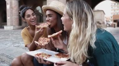 Üç neşeli çok ırklı kadın sokakta pizza yiyor.