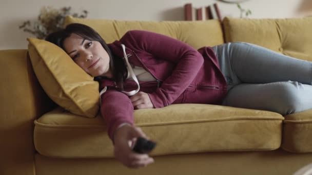 Znudzona młoda kobieta ogląda telewizję leżącą na kanapie w domu — Wideo stockowe