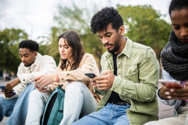 Diverse young students friends using smart phone while sitting in city street