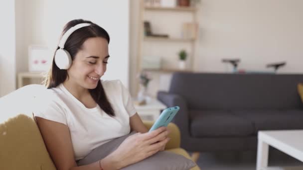 Feliz mujer adulta joven positiva usando el teléfono mientras escucha música en casa — Vídeo de stock