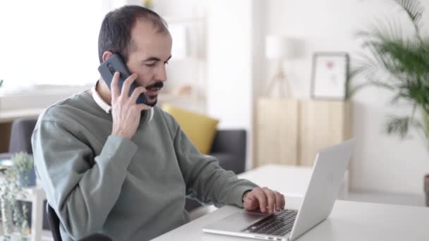Mid vuxen man pratar på mobiltelefon medan du arbetar på bärbar dator från hemmakontoret — Stockvideo