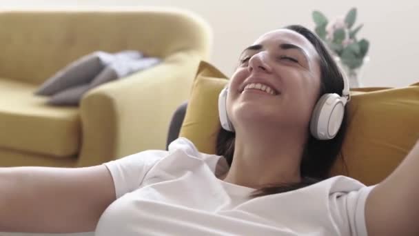 Glada ung kvinna dansar medan du lyssnar på musik som ligger på soffan — Stockvideo