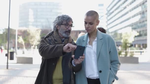 Zwei Geschäftsleute gehen und reden über die Arbeit, während sie ihr Smartphone checken — Stockvideo