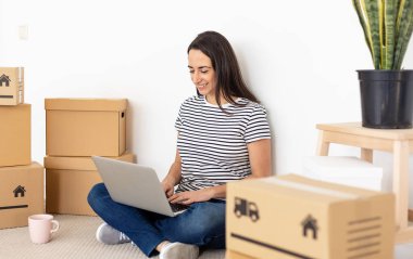 Yeni apartman dairesinde dizüstü bilgisayar kullanan gülümseyen genç kadın. 