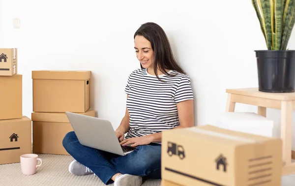 Χαμογελώντας νεαρή γυναίκα χρησιμοποιώντας φορητό υπολογιστή σε νέο διαμέρισμα ιδιοκτησίας — Φωτογραφία Αρχείου
