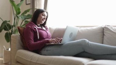 Genç bir kadın dizüstü bilgisayar kullanırken koltukta dinleniyor.