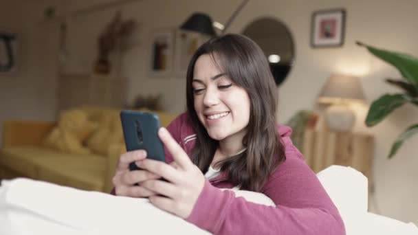 Mooie jonge volwassen vrouw ontspannen thuis met behulp van mobiele telefoon zitten op de bank — Stockvideo