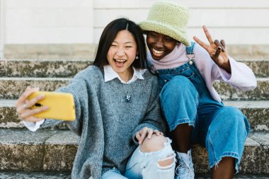 İki çok ırklı genç kadın cep telefonuyla selfie çekerek eğleniyor.