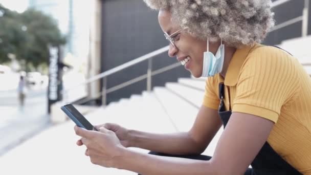 Heureuse jeune femme hispanique avec des cheveux afro en utilisant un téléphone mobile à l'extérieur — Video