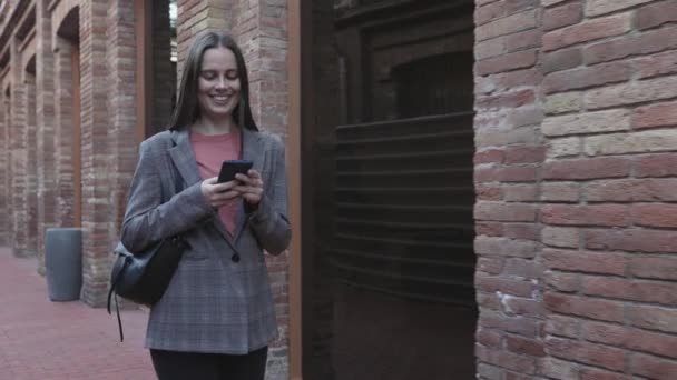 Sourire jeune femme adulte marchant dans la rue de la ville tout en utilisant un téléphone mobile — Video