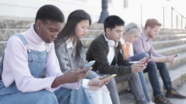 Multiracial jeunes amis se détendre avec des téléphones mobiles assis à l'extérieur — Video