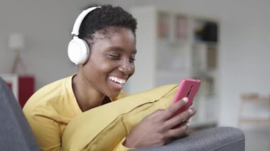 Evdeki kanepede yatarken akıllı telefon kullanan mutlu siyah kadın.