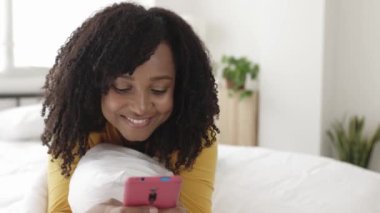 Rahat, güzel, Afro-Amerikalı bir kadın. Yatakta cep telefonu kullanıyor.