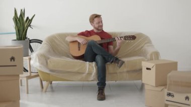 Neşeli genç adam yeni evinde gitar çalıyor.
