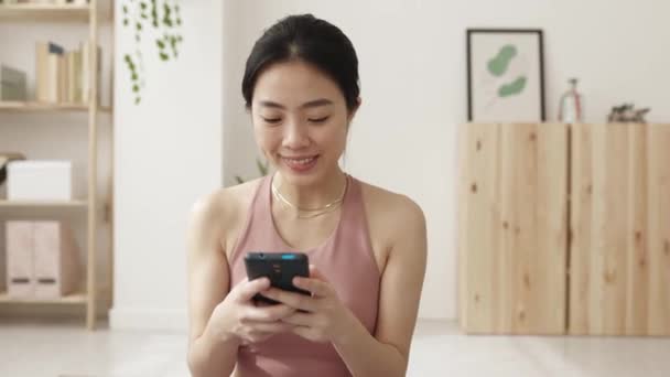 Joven mujer asiática usando el teléfono móvil después del entrenamiento rutina de ejercicios en casa — Vídeo de stock