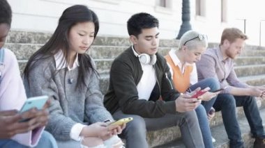 Açık havada oturan akıllı telefonlarla rahatlayan çok ırklı gençler