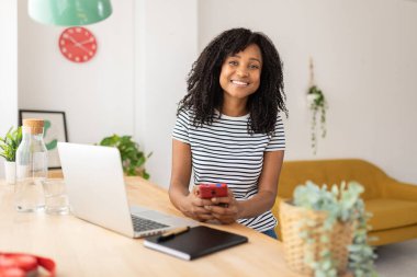 Evde masaüstünde çalışırken akıllı bir cep telefonu kullanan gülümseyen kadın.