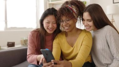 Three multiracial women friends having fun using mobile phone at home