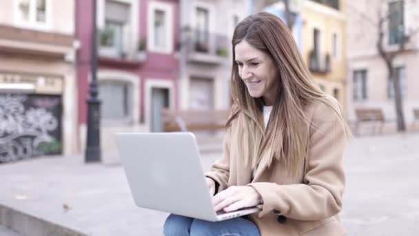 Young adult woman using laptop computer while sitting outdoors in city street — Stok video