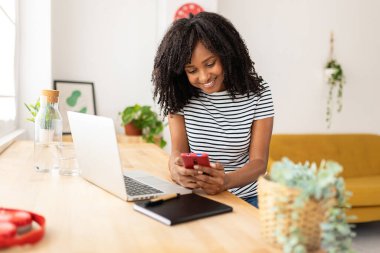 Genç İspanyol asıllı Amerikalı kadın evde dizüstü bilgisayarla çalışırken cep telefonu kullanıyor.