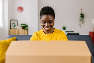 Mutlu Afro-Amerikalı kadın çevrimiçi alışveriş yaptıktan sonra paketleri açıyor, gülümseyen kadın paketleri alıyor ve evdeki koltukta otururken açıyor.