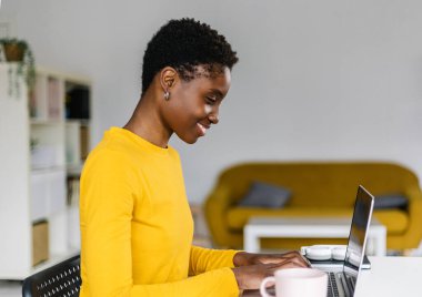 Ev ofisinden dizüstü bilgisayarla çalışan gülümseyen Afrikalı bir kadın. Çalışma masasında otururken online çalışan serbest çalışan bir kadın.