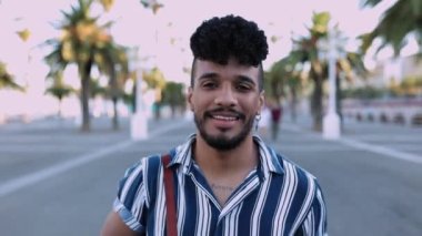 Yazın kameraya gülümserken dışarıda dolaşan kendine güvenen genç, modaya uygun Latin bir adamın ön görüntüsü. Yüksek kalite 4k görüntü